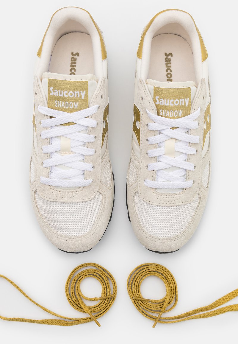 Un paio di sneaker Saucony Shadow beige e oro con lacci bianchi annodati, accompagnati da lacci d'oro a spirale separati su sfondo bianco.