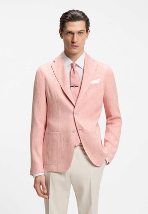 Man wearing light pink blazer, white shirt, matching pink tie with tie clip, beige trousers, posing against white background.