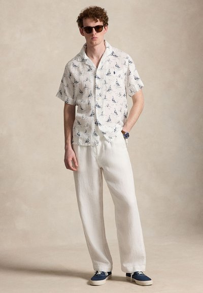 Homme portant des lunettes de soleil, chemise blanche à manches courtes avec imprimé de voilier, pantalon blanc, baskets bleues, debout devant un fond neutre.