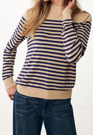 Mujer con suéter de rayas horizontales beige y azul marino y vaqueros azul oscuro, con la mano apoyada cerca del cuello y la cadera.