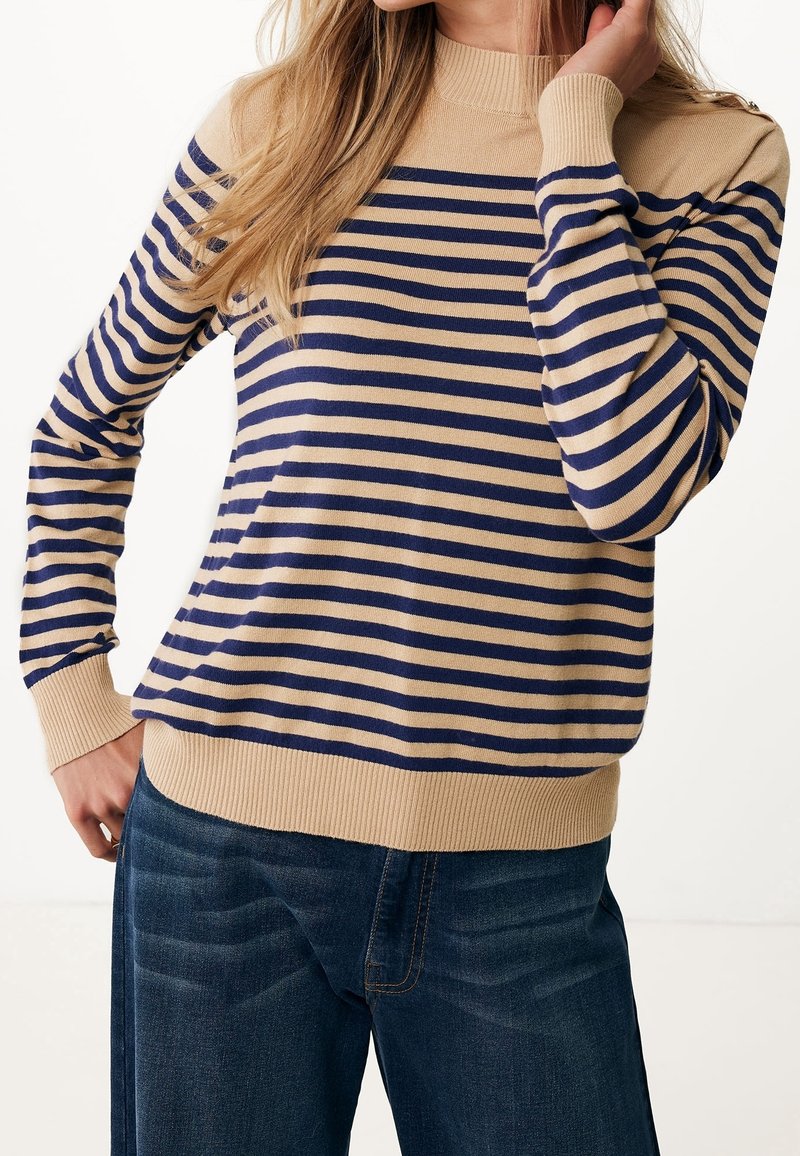 Femme portant un pull à rayures horizontales beige et bleu marine et un jean bleu foncé, la main reposant près du cou et de la hanche.