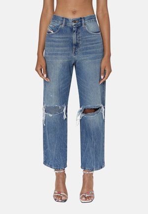 Kobieta ubrana w niebieskie, wysokowalne jeansy o prostych nogawkach z przecieranymi dziurami na kolanach i postrzępionymi krawędziami oraz srebrne sandały na obcasie z paskami wokół kostek.