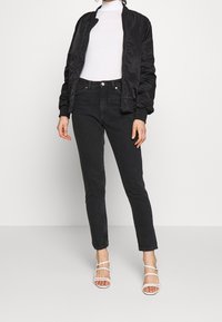 Chaqueta bomber negra sobre un suéter de cuello alto blanco, jeans pitillo negros y sandalias blancas con tiras. Texturas suaves y diseño ajustado.