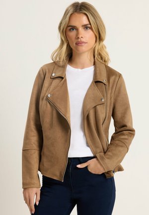 Kvinde med blondt hår iført tan ruskind jakke over hvid skjorte og mørke jeans, står med den ene hånd i lommen mod en lys baggrund.