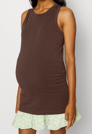 Zwangere persoon draagt een mouwloze bruine zwangerschaps top en een lichtgroene bloemenprint rok met ruches, staat met ontspannen handen langs het lichaam.