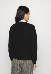 Schwarzer Strickpullover mit geripptem Muster, Rundhalsausschnitt und langen Ärmeln, getragen über karierten Hosen. Die Rückansicht zeigt minimale Details.