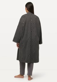 Schwarz-weißer gemusterter Kimono mit geschwungenen Linien, langen Ärmeln und geradem Schnitt, über grauen Hosen getragen, von hinten gezeigt.