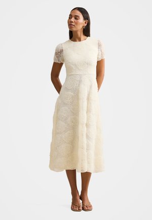 Vrouw die een crèmekleurige midi-jurk van kant draagt met korte doorschijnende mouwen, staand met de handen achter de rug en beige sandalen aan.