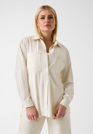 Frau mit glatten blonden Haaren, die ein übergroßes cremefarbenes Hemd mit Knöpfen über passenden Hosen vor einem schlichten weißen Hintergrund trägt.