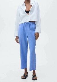 Bílá zkrácená košile s dlouhými rukávy, v kombinaci s světle modrými kalhotami na šnůrku. Kalhoty mají rovný střih a hladkou texturu. Černé sandály.