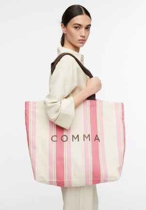 Donna in outfit color crema che porta una grande borsa a righe rosa e crema con manici scuri e scritta "COMMA" sul davanti.