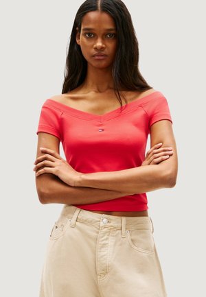 Donna con lunghi capelli scuri che indossa una maglia rossa a spalle scoperte e pantaloni beige, in piedi con le braccia incrociate davanti a uno sfondo neutro.