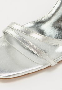 Sølvmetallisk blokk-hæl sandal med to tynne overlappende stropper, blank overflate og firkantet tå design.