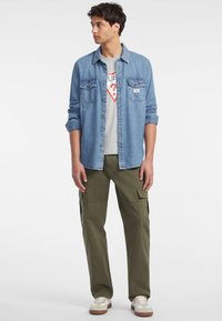 Camisa de mezclilla azul claro con dos bolsillos en el pecho, camiseta gráfica gris debajo, pantalones cargo oliva y zapatillas blancas con detalles en marrón.
