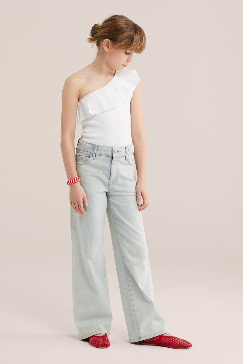 Mladý človek stojaci, oblečený v bielom topu na jedno rameno, svetlomodré široké nohavice, červený náramok a červené pletené balerínky na béžovom pozadí.