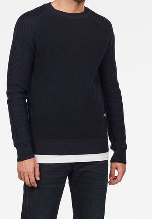 Suéter de punto azul marino con mangas texturizadas, cuello redondo y puños acanalados. Lleva puesto sobre una camiseta blanca, combinado con jeans oscuros.