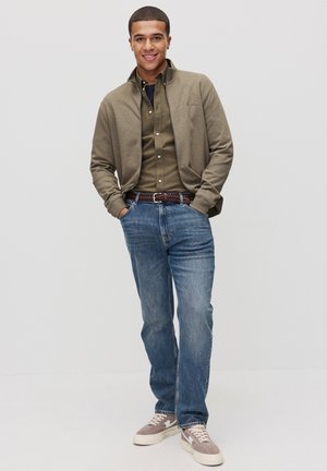 Afslappet outfit med en lys brun zip-up jakke over en mørkegrøn skjorte med knapper, parret med blå jeans og grå sneakers.