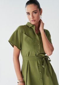 Abito camicia verde oliva con maniche corte, chiusura con bottoni davanti e vita annodata. Presenta tasche frontali superiori e un tessuto texturizzato.