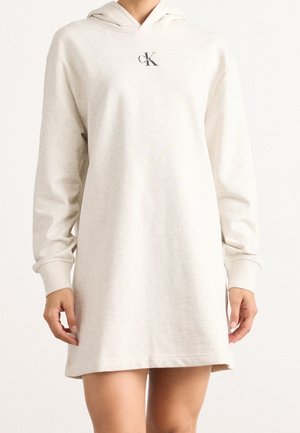Woman wearing a cream hooded long-sleeve sweatshirt dress with a small black "cK" logo on the chest.