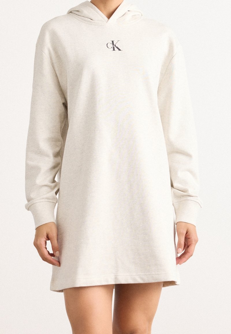 Femme portant une robe sweat à capuche crème à manches longues avec un petit logo noir « cK » sur la poitrine.
