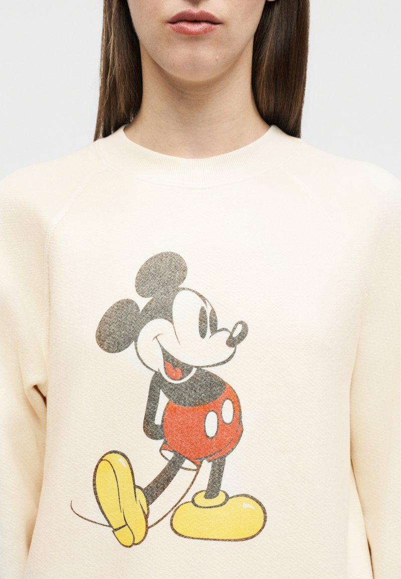 Femme portant un sweat-shirt crème avec un motif vintage de Mickey Mouse en short rouge et chaussures jaunes.