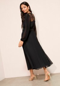 Fekete, plisséelt ruha csipkés hátsó résszel, hosszú ujjakkal és megkötött derék részsel, arany, pántos magassarkú szandálokkal párosítva.