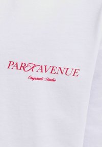 Λευκό ύφασμα με κόκκινο κείμενο "PART AVENUE" και μικρότερο κείμενο με πλάγια γραφή "Originals Studio" τυπωμένα πάνω του.