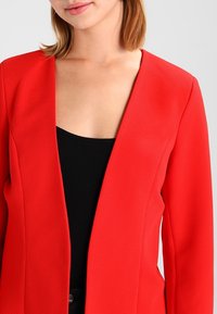 Červený blazer s texturovanou tkaninou, otevřeným designem vpředu a dlouhými rukávy. Nosit se přes černý top, předvádějící štíhlý střih a moderní styl.