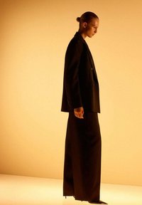 Traje negro a medida con pantalones anchos y blazer cruzado. Tejido liso, diseño minimalista, sobre un fondo amarillo cálido.