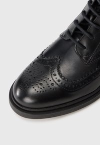 Black leather brogue shoe with perforated detailing and black laces on a white background, showing the toe and part of the sole.