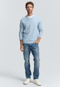 Svetlo modra pulover z v-izrezom, mehka tekstura, kombiniran s svetlo modrimi kavbojkami in belimi supergami, stoji na nevtralnem ozadju.