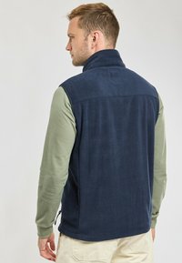 Gilet en polaire marine avec col haut, poches zippées et texture douce. Porté par-dessus une chemise verte à manches longues, présentant une coupe droite.