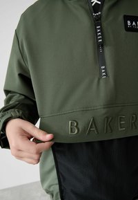 Giacca con pullover verde e mezzi zip, rete nera testurizzata nella parte inferiore, e logo "BAKER" impresso sul davanti.