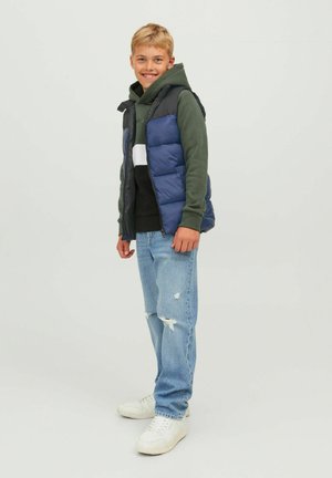 Gilet imbottito blu navy e grigio sopra un hoodie verde e una maglietta nera, abbinato a jeans strappati blu chiaro e sneakers bianche. Outfit casual.