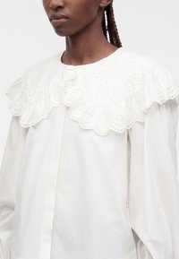 Femme noire portant un chemisier blanc avec un grand col en dentelle ajourée et de longues manches bouffantes, visible de la poitrine au cou.