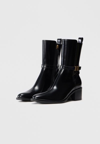 Par de elegantes botas de cuero negro hasta la mitad de la pantorrilla, con puntas afiladas, tacones rectangulares, cremalleras laterales y correas con hebillas doradas sobre un fondo blanco liso.