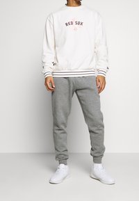 Felpa bianca con ricamo "RED SOX" e dettagli bordeaux, abbinata a pantaloni della tuta grigi e sneaker bianche. Abbigliamento sportivo casual.