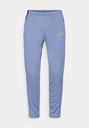 Svetlo modri športni hlače iz mehkega blaga, ki imajo črne stranske panele, elastični pas in rumen Nike logotip na levem stegnu.