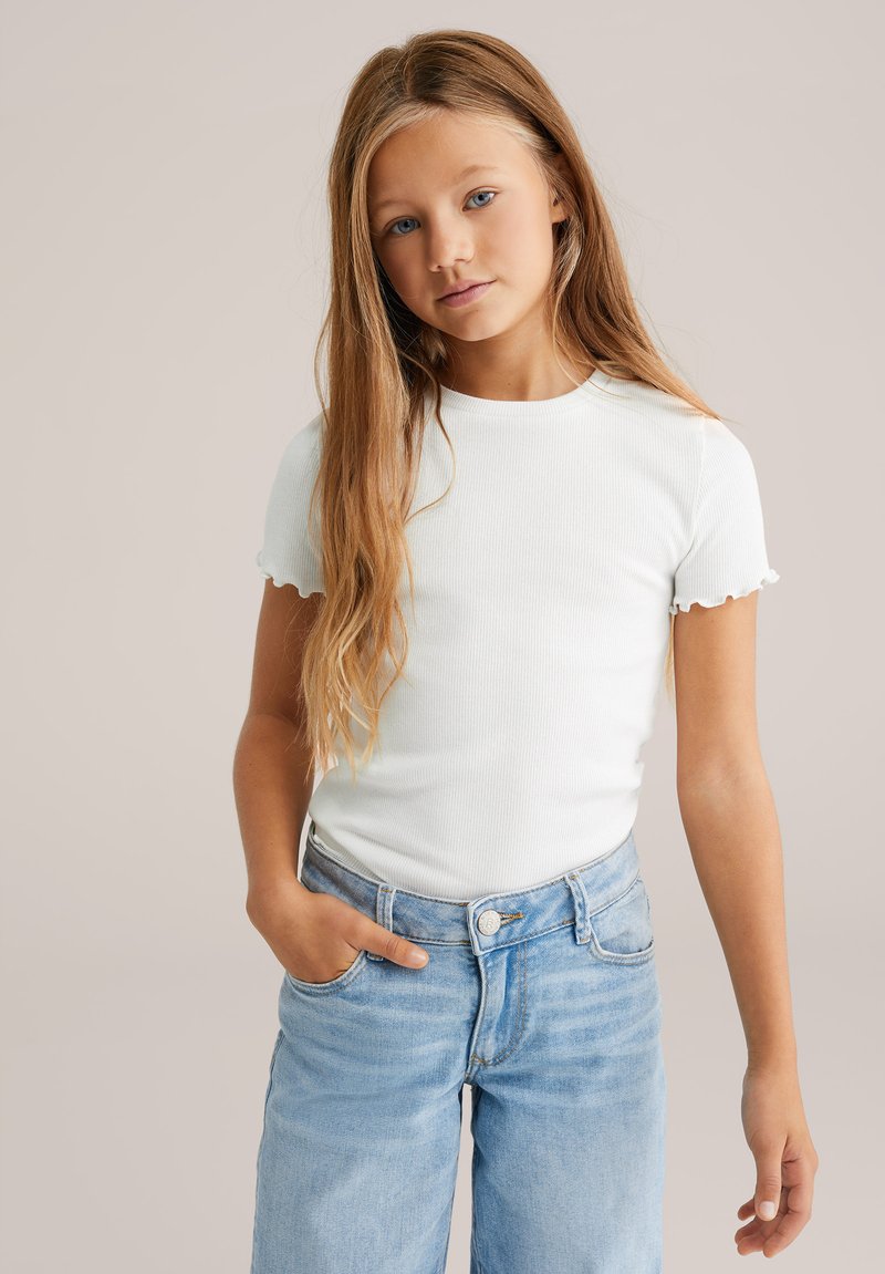 Biała, żebrowana koszulka z krótkimi, falbaniastymi rękawami; połączona z jasnoniebieskimi dżinsami z wysokim stanem, posiadającymi zapięcie na guzik i kieszenie.