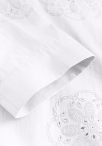 Biała tkanina z dokładnym haftem kwiatowym w ton w ton. Mankiet jest starannie wykończony czystymi szwami.