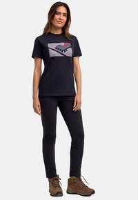 Schwarzes Baumwoll-T-Shirt mit großem grafischem Aufdruck in Grau, Rot und Schwarz. Kombiniert mit dunkler Hose und braunen Wanderschuhen.