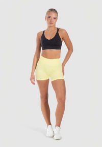 Sort sports-bh med rynkedetalje, kombineret med shorts i lys gul med høj talje. Modellen har hvide sneakers på; ensfarvet lysegrå baggrund.