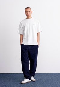 T-shirt blanche à manches courtes avec un texte bleu, associé à un pantalon bleu marine comportant des poches et des baskets blanches sur un fond de tapis bleu.