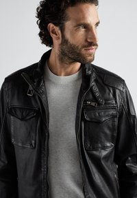 Schwarze Lederjacke mit Reißverschluss- und Klappentaschen, Knopfverschlusskragen und einem strukturierten grauen Pullover darunter.