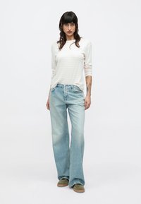 Chemise gris clair à manches longues avec des rayures subtiles, associée à un jean évasé bleu clair. Les chaussures sont vert olive avec une semelle en caoutchouc.