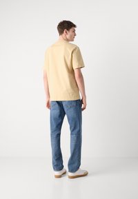 Světle žlutá košile s krátkým rukávem umístěná na pozadí, v kombinaci se světle modrými džíny. Model má na sobě bílé tenisky. Minimalistický design.