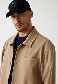 Giacca beige con colletto a scollatura e una tasca sul petto, indossata sopra una camicia blu scuro, abbinata a un berretto blu con logo.