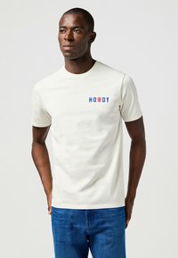 Camiseta de algodón crema con la palabra "HOWDY" en texto rojo y azul en el lado izquierdo del pecho. Mangas cortas y cuello redondo.