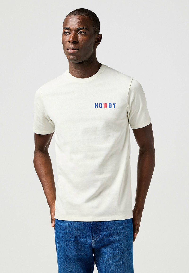Camiseta de algodón crema con la palabra "HOWDY" en texto rojo y azul en el lado izquierdo del pecho. Mangas cortas y cuello redondo.