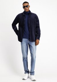 Camicia di velluto a coste blu marina sopra un dolcevita blu marina, abbinata a jeans azzurri e sneaker bianche, con vestibilità rilassata e due tasche sul petto.